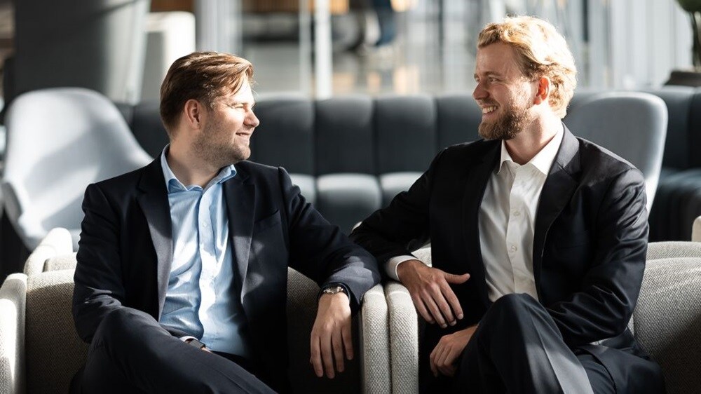 Two people seated in armchairs in a modern office lounge, engaged in a conversation with an open workspace visible in the background.