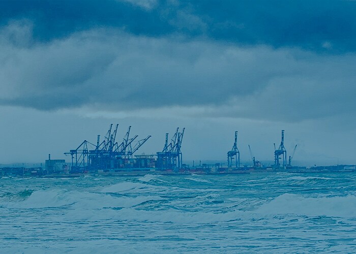 Stormy ocean waves with a view of a port featuring multiple container cranes under cloudy skies