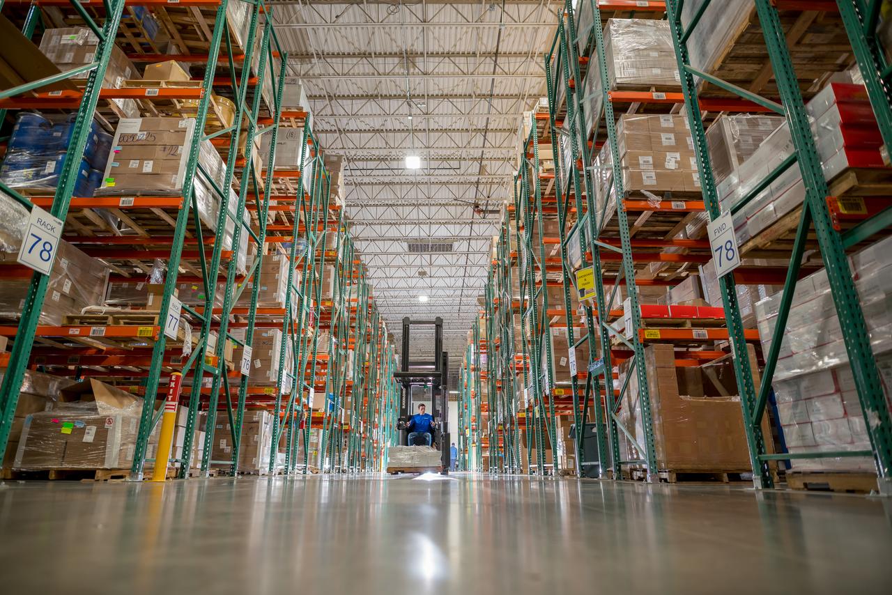 A man in blue overalls drives a forklift down the aisle of a warehouse. On either side the shelves reach up to the ceiling and are stacked with packages.