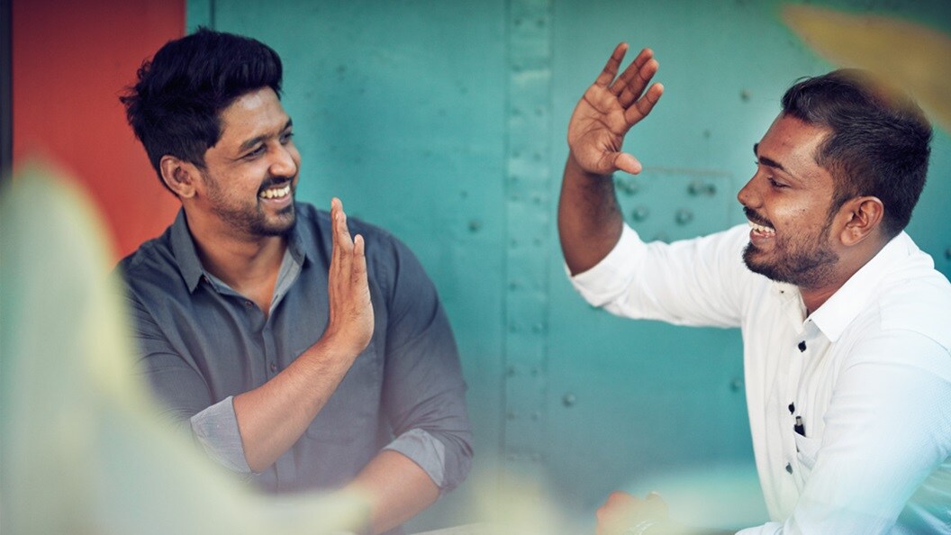 Two men with dark hair and brown skin smile and high five with a blue shipping container in the background. 