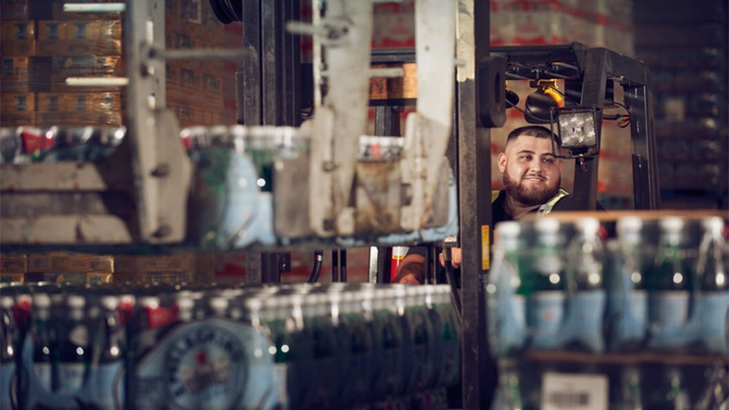 A smiling man with short dark hair and a beard drives a forklift. In the foreground are several palettes with water bottles.