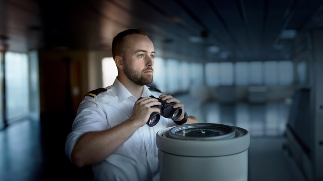 Ship officer in a white uniform holding binoculars, looking ahead on a vessel's bridge.