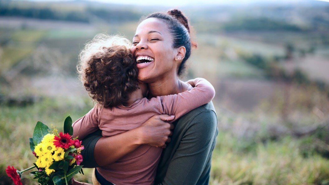 A woman and her child embrace warmly in a sunlit field, surrounded by tall grass and wildflowers.