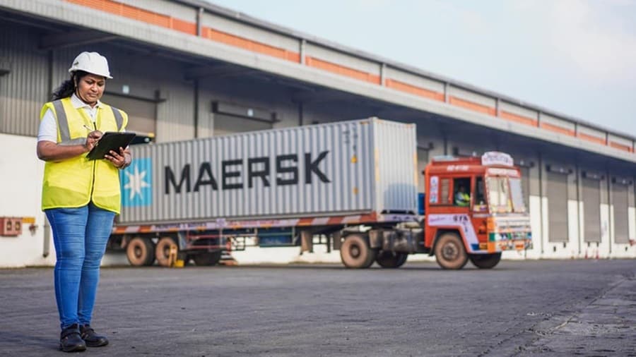 A woman with a hard hat and safety vest stands in front of a warehouse, indicating her involvement in the construction industry.