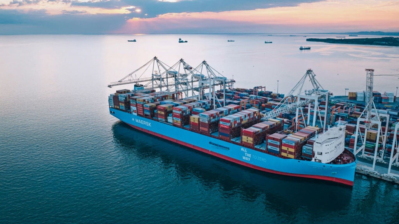A large container ship is docked at a busy port, with cranes and cargo containers visible in the background.