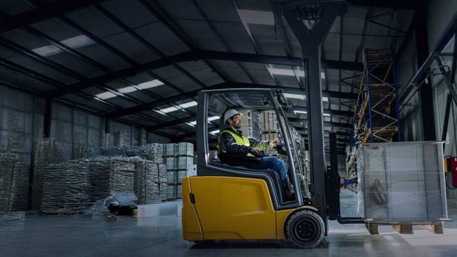 A warehouse scene featuring a man driving a forklift, maneuvering between storage racks filled with goods.