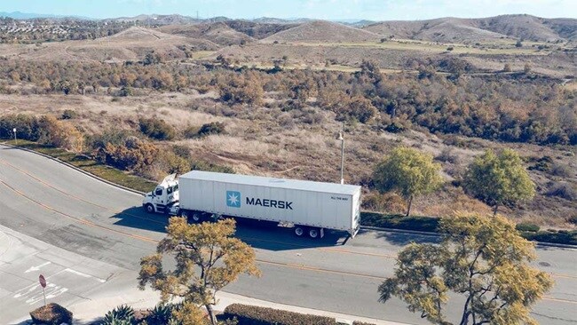 A Maersk container truck is driving on a road surrounded by trees and hills.