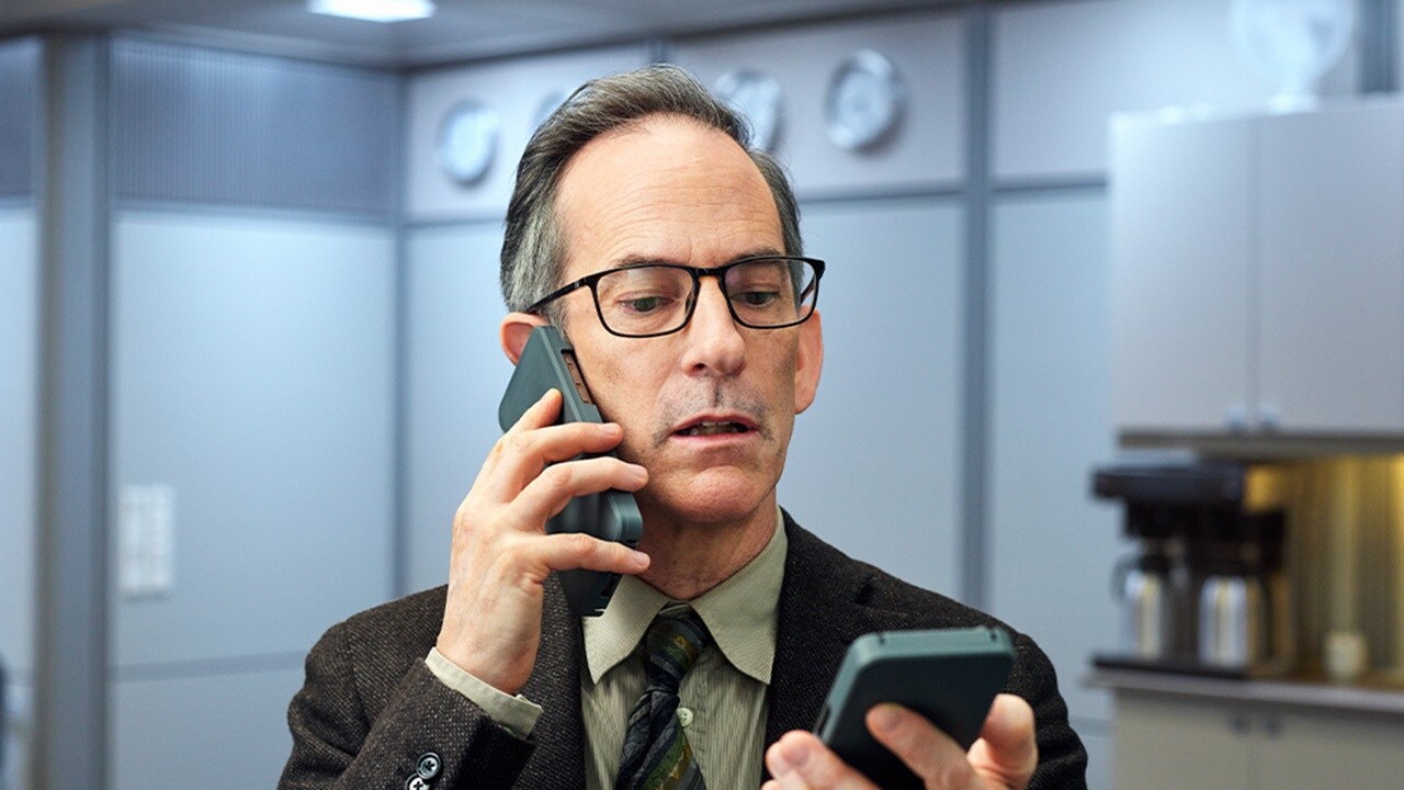 A man in a suit speaks on a phone while holding a smartphone, surrounded by a modern office setting.